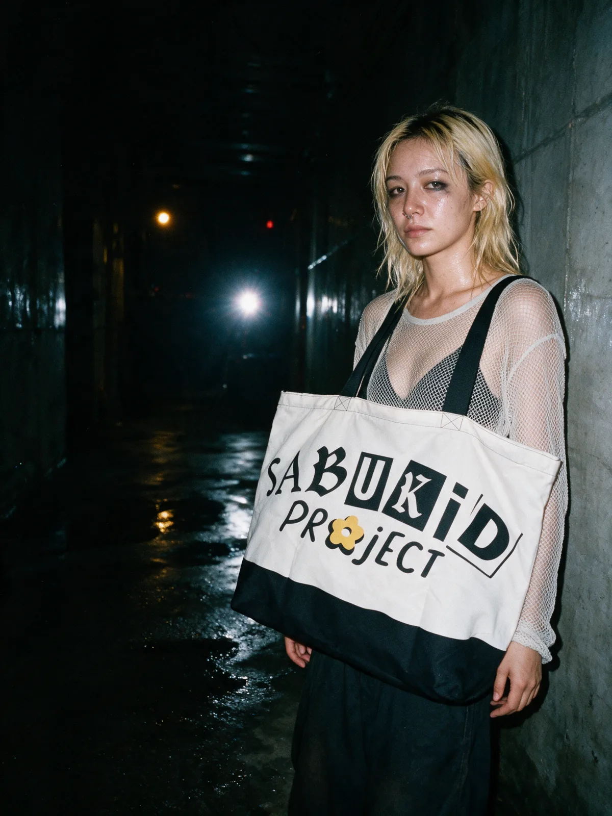 A woman in a mesh top against a concrete wall at night, the bone tote on her shoulder, lit by direct flash.