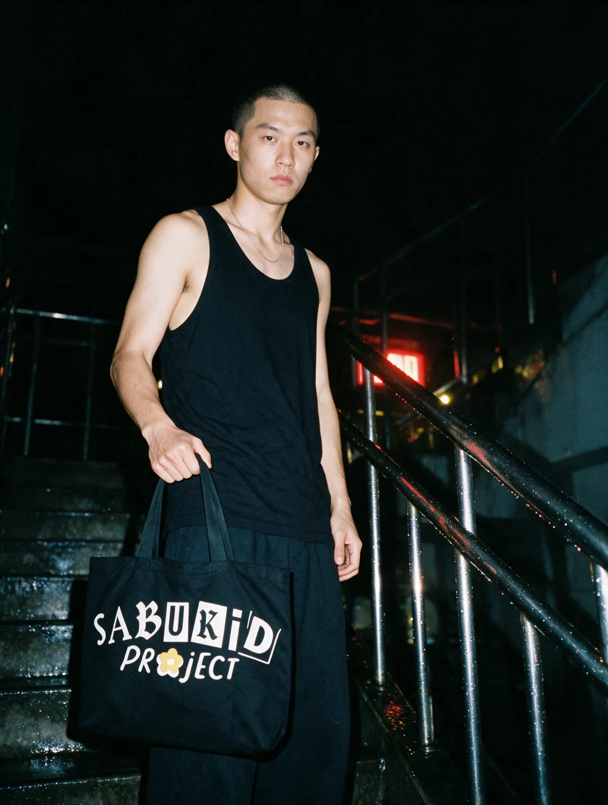 A man in a black tank top on a wet stairwell under a red exit sign, holding the black tote.