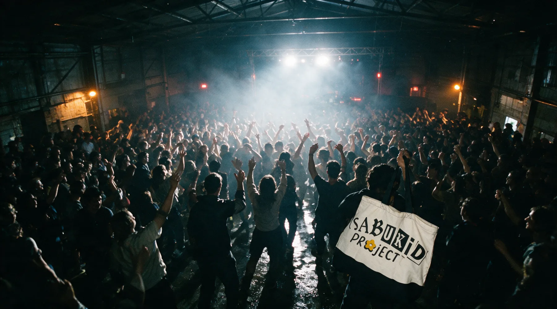 A warehouse floor packed with people in haze, lit from behind, arms raised. One bone tote held up at the right of the crowd.