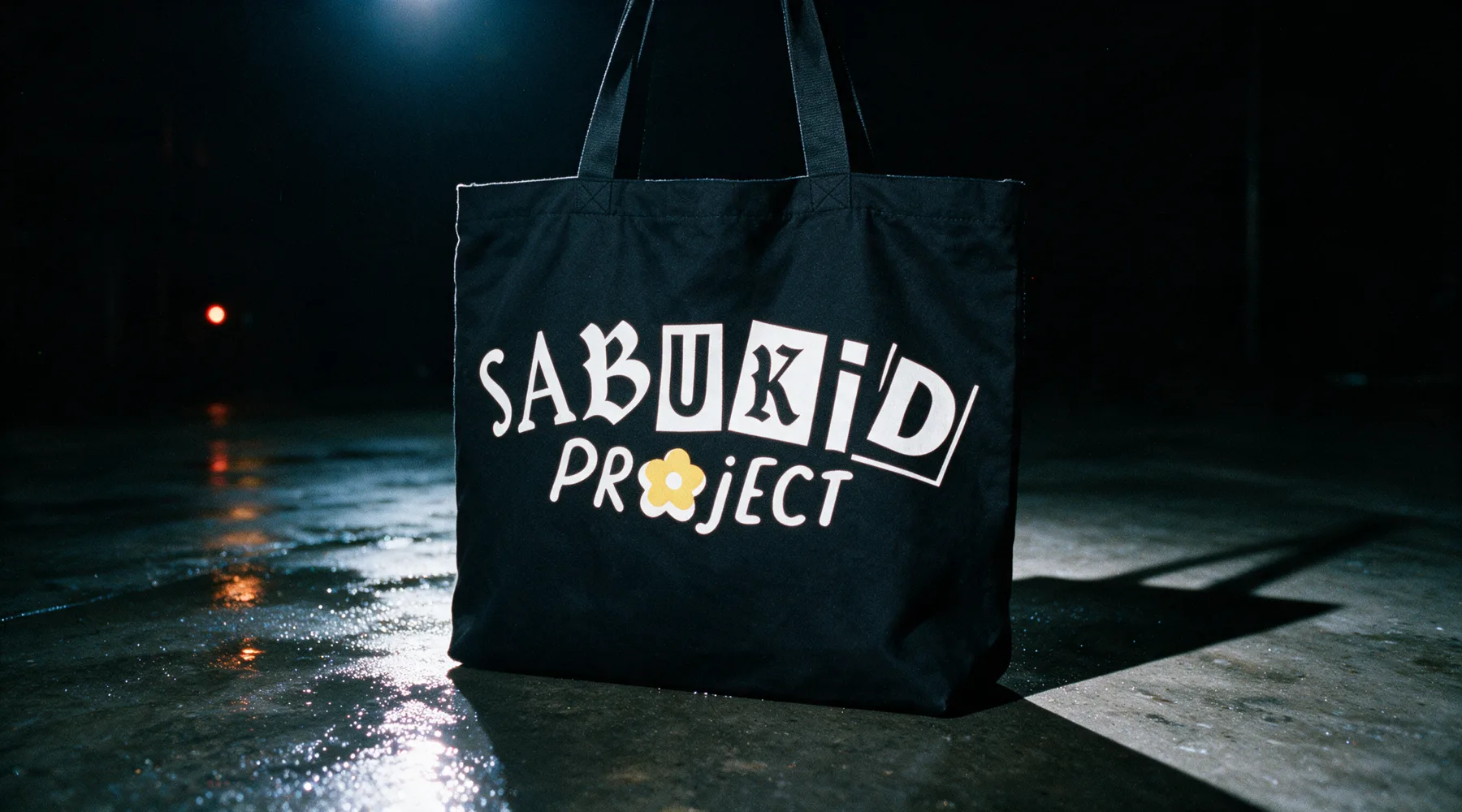 The all-black tote standing alone on wet concrete under one hard light, the mark printed in bone white with the yellow flower.